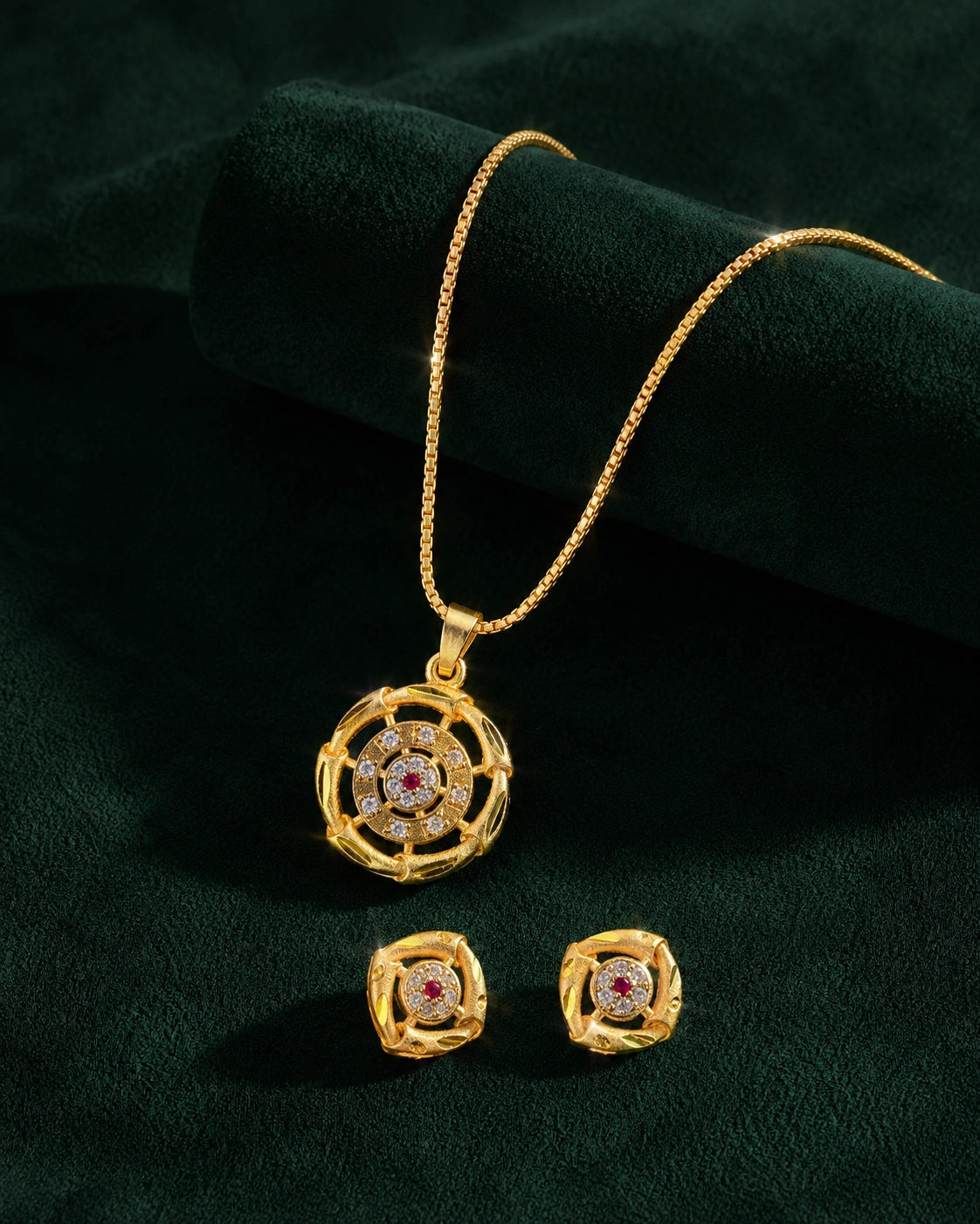 Gold jewelry set featuring a round pendant necklace and matching stud earrings with a central red gemstone and clear stones, displayed on a dark green velvet background.