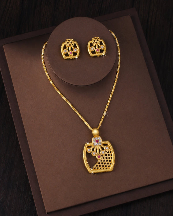 Gold jewelry set featuring a necklace and matching earrings displayed on a brown presentation box. The pendant and earrings have an ornate openwork design with small pink gemstone accents.