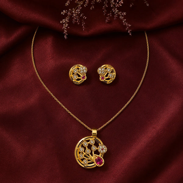 Gold jewelry set featuring a pendant necklace and matching stud earrings, decorated with clear stones and a red teardrop gemstone, displayed on a draped dark red satin fabric.