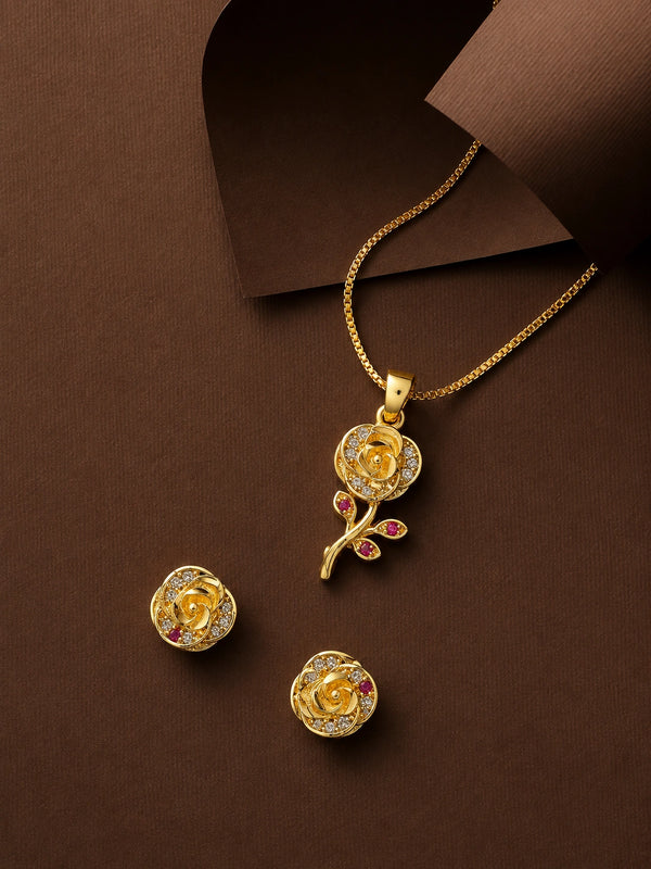 Gold rose pendant necklace and matching stud earrings with small pink gemstone accents, arranged on a dark brown textured background.