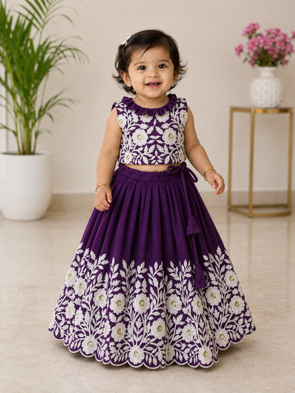 Child wearing a purple floral dress standing indoors with a plant and table in the background.